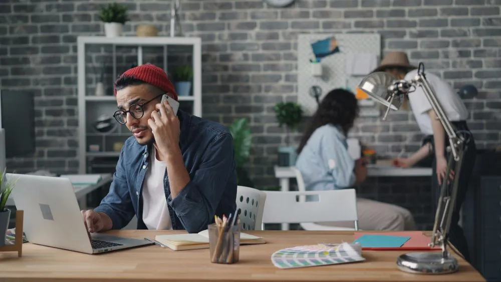 Hombre trabajando en una oficina moderna, hablando por teléfono mientras usa una laptop